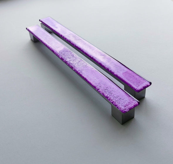Set of two sparkly bright purple fired glass fridge pulls made of long glass sticks attached over a silvery metal base. Slightly textured glass furniture handles with rounded edges.