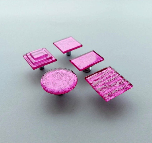 Fired glass cabinet knobs in bright pink made of round, square and rectangular glass pieces attached over a metal base. Slightly textured glass furniture handles with rounded edges.